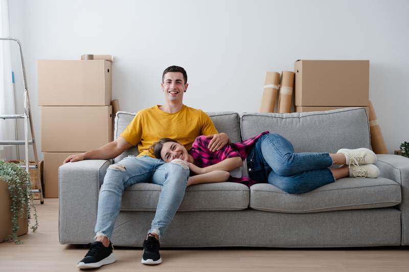 A man and a woman relax on a sofa surrounded by moving boxes in an empty room.