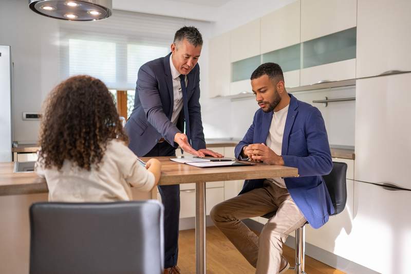 Agent reviews title insurance documents with man and woman at the kitchen table of an empty home.